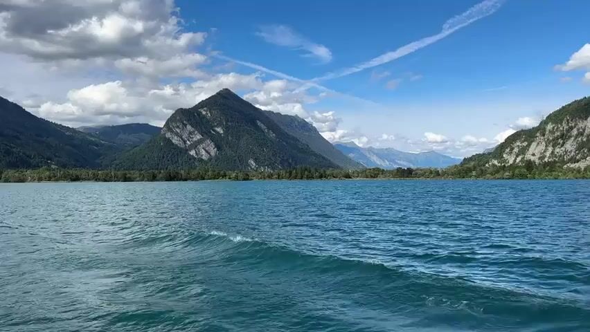 This video shows a wide shot of a lake with mountains in the background. The camera is positioned low to the water, giving the impression of being on a boat. The water is a vibrant blue and has ripples from movement. In the distance, there are lush green mountains dotted with trees. The sky is a clear blue with scattered white clouds, some of which appear as contrails. The mountains are layered, with some appearing closer and more detailed than others in the far distance. The scene is bright and appears to be a sunny day. There are no people or distinct objects visible in the video, only the natural landscape. The video captures the movement of the water and the expansive view of the lake and mountains.