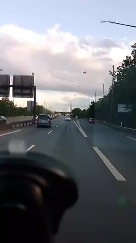 The video shows a forward-facing view from inside a vehicle traveling on a multi-lane highway under a cloudy sky. Several cars and a red truck are visible on the highway, with traffic appearing to be moderate. The road is paved with asphalt and marked with white lines. On the left side of the highway, there is a barrier and vegetation, as well as some buildings in the distance. On the right side, there is more vegetation and trees. The sky is filled with large, fluffy clouds, and the overall lighting suggests it is either early morning or late afternoon. The lower part of the frame shows the interior of the vehicle, specifically what appears to be the dashboard and possibly the steering wheel. There are no visible people outside the vehicles.