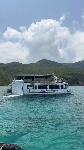 A large, white double-deck boat named "Ocean World" and "Crazy Party Hopping" is anchored or moving slowly in clear, turquoise water under a partly cloudy sky. The boat features a white slide-like structure at its stern, and red flags are visible on its upper deck. Several people can be seen on board, suggesting a party or tour event. In the background, lush green mountains rise from the coastline, indicating a scenic natural setting. Visible text on the boat includes "OCEAN WORLD" in yellow and "Crazy Party Hopping" in colorful letters. Other details like "KB-1088" and "TY 99K" are also present on the boat's side. The scene depicts a vibrant daytime atmosphere in a coastal area, likely a natural heritage site in Trang, Vietnam.