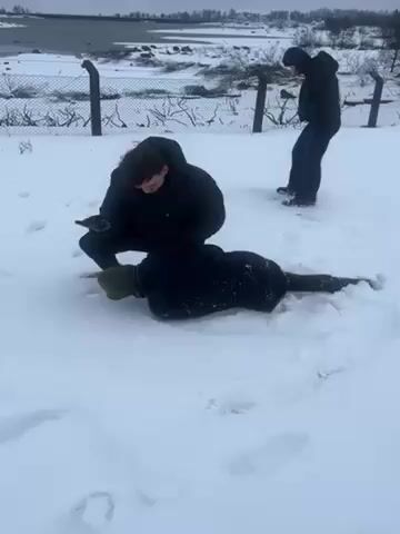 Two men are outside in a snow-covered field. One man is on top of another man who is lying face down in the snow. The man on top is wearing a black coat and dark pants. He appears to be holding the man on the ground. The man on the ground is wearing a black coat and a beanie. A third person is standing in the background wearing dark clothing. There is a wire fence behind the men. In the distance, there is a body of water and bare trees. It appears to be a cold, winter day.