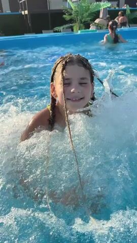 A young girl with braided hair is playing in a pool. She is in the center of the frame, splashing water around her. Her eyes are closed and she appears to be enjoying herself. In the background, other people are in the pool and on the edge of the pool. There are lounge chairs and umbrellas visible in the background, suggesting a recreational area. The water is blue and clear, and the sun is shining.