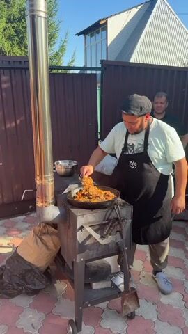 A man wearing a black apron over a light blue t-shirt and a dark cap is standing outdoors in front of a metal stove. He is stirring food in a large, black skillet with a spatula. The food appears to be a mixture of rice, meat, and vegetables. There is a silver bowl to the left of the skillet and a metal chimney pipe extending upwards from the stove. Behind the man, another man is partially visible, looking towards the skillet. The background consists of a brown wooden fence and a building. The ground is paved with interlocking tiles. A large brown bag is placed on the ground to the left of the stove. The lighting suggests it is daytime, possibly late afternoon due to the long shadows.
