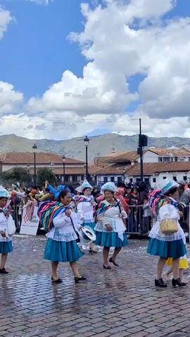A group of women and girls dance in a parade on a cobblestone street in Cusco, Peru. They wear matching white blouses, teal skirts with white lace trim, colorful shawls, and blue hats. Some of the women carry small white bags. The dancers move toward the right side of the frame. They are surrounded by a crowd of onlookers behind metal barricades. Buildings, including churches, are visible in the background. Mountains are visible in the distance under a partially cloudy sky. Confetti litters the street.