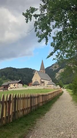 This is a vertical, eye-level video clip of a scenic path leading towards a picturesque village in the Italian Alps. The path is made of gravel and lined with a rustic wooden fence, with lush green grass growing on the other side. In the distance, a charming village with traditional alpine architecture is nestled among rolling green hills and majestic mountains. A prominent church with a tall, pointed steeple stands at the heart of the village, its stone facade weathered and timeless. The sky above is a mix of blue and white clouds, suggesting pleasant weather, possibly late afternoon given the soft lighting. A few people can be seen walking along the path in the distance, adding a sense of peaceful activity to the tranquil setting. The overall mood is serene and idyllic, capturing the beauty of rural Italy. There is no visible text in the media file.