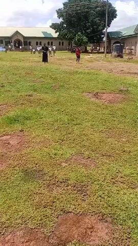 The video shows a partially grassy and partly muddy field with a building in the background. There are several people visible in the distance, some walking and some standing near the building. A person in the foreground wearing a white t-shirt with "NYSC" printed on the back, green pants, a green cap, and brown shoes, is walking across the field. This person is also holding a phone in their hand and appears to be recording the video. The weather looks sunny with a cloudy sky.