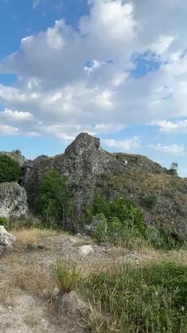 The video shows a panorama of a rural landscape under a partly cloudy sky. In the foreground, there is rough terrain with dry grass and rocks. The middle ground features a rocky hill with sparse vegetation, leading to a view of a village nestled in a valley. The village has houses with red roofs, surrounded by green trees. In the background, rolling hills and mountains stretch towards the horizon. The sky is blue with white and gray clouds, and sunlight streams through gaps in the clouds. The camera pans slowly across the scene, revealing the vastness of the landscape.