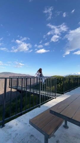 A young woman stands on a metal viewing platform that extends over a forested gorge. The sky is blue with puffy white clouds. Mountains are visible in the distance. The woman straightens up from leaning over the railing, smiles, and walks across the platform. A picnic table is visible in the lower right foreground.