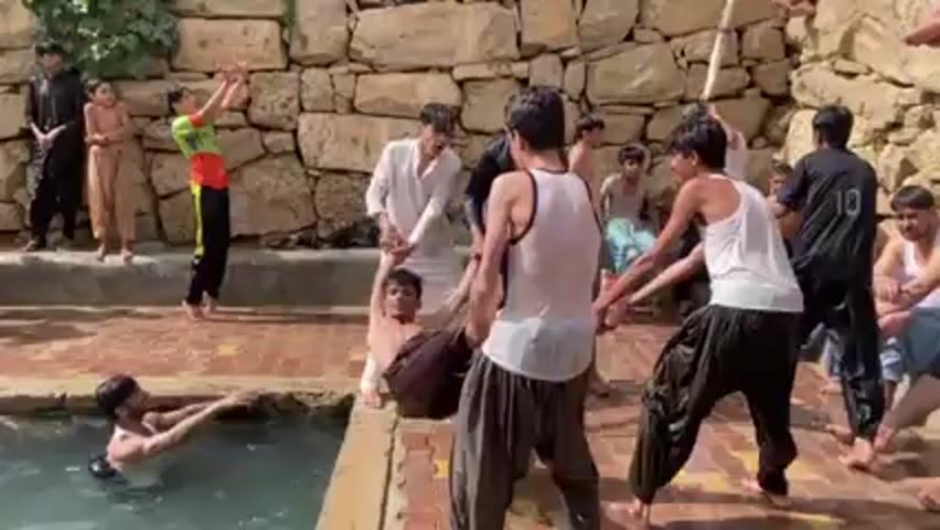A group of men and boys are gathered at a pool of water, some in the water, some on the edge. There is a stone wall behind them. Some of the men are grabbing at each other, apparently playfully, and throw another man into the pool. Everyone is laughing.