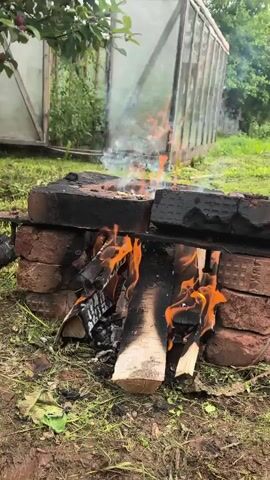 A fire burns in an outdoor setting. The fire is contained within a makeshift structure of bricks. Several logs are burning, with flames and smoke visible. In the background, a greenhouse or similar structure can be seen, along with greenery and grass. The overall impression is of a rural or garden environment where a fire has been lit.