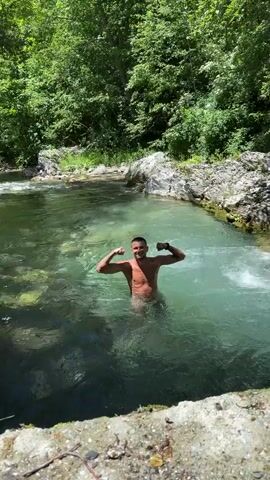 A man is standing in clear, shallow water with his arms flexed and smiling at the camera. He is surrounded by rocks and lush green trees, indicating he is in a natural outdoor setting. The water appears to be a river or a pool within a river. The man is shirtless.