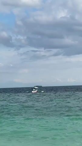 The video shows a wide shot of the ocean under a cloudy sky. The water in the foreground is a vibrant turquoise, transitioning to a darker blue further out. A small white boat with a canopy is visible in the middle distance, moving from left to right. There appear to be a few people on the boat. In the background, land or islands can be seen on the horizon, with faint mountain shapes. Another, smaller white boat is visible to the left of the main boat, also moving. The waves are moderate, creating a shimmering effect on the water's surface. The overall scene is calm and serene, suggesting a pleasant day at sea.