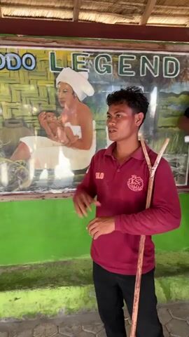 A man wearing a maroon polo shirt and black pants stands in front of a mural. He is holding a wooden stick in his right hand and has his left hand raised. The mural depicts a woman holding a baby, a Komodo dragon, and a mountainous landscape. The man appears to be speaking or giving instructions. The environment looks like an outdoor setting with a thatched roof overhead.