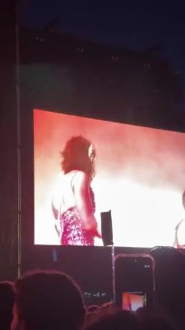 The video shows an outdoor concert, likely at night, viewed from the audience. A large screen displays two performers. One is singing into a microphone, wearing a sparkly outfit. The other performer is wearing a sparkly outfit and a monkey mask. The screen is lit with red light. In the foreground, audience members hold up phones, recording the performance. The camera slightly shakes.