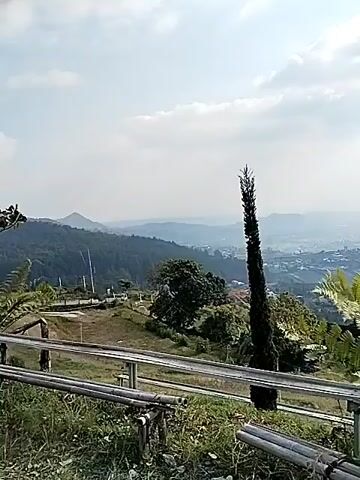 A high-angle shot shows a mountainous landscape under a cloudy sky. In the foreground, a wooden bench is partially visible, along with some bamboo poles and green foliage. A tall, slender evergreen tree stands prominently in the mid-ground, to the left of center. Further back, lush green trees and ferns are scattered across rolling hills. In the distance, a hazy valley unfolds, revealing a town or city with buildings nestled amongst the terrain. The overall scene is natural and serene, with a hint of human habitation in the distance. The video appears to be a panning shot, moving from left to right.
