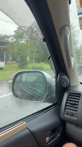 The video shows the interior of a vehicle driving on a road during heavy rain. The view is from the passenger seat, looking out the windshield and side window. Raindrops are visible on the windshield, and the road ahead is wet and reflects the overcast sky. Trees and vegetation line the sides of the road. The dashboard and part of the car's interior are visible in the foreground. There are also stickers on the inside of the windshield. The scene appears to be in a rural or suburban area.