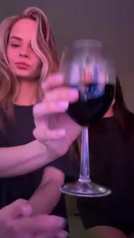 Two women are posing for a video, each holding a glass of red wine. The woman on the left has blonde hair parted to the side. The woman on the right has long brown hair. Both are wearing black t-shirts. The background is a room with a couch and a chandelier. The video is lit with pink and purple light. They both make kissy faces at the camera.