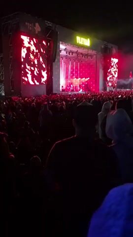 This is a video of a large outdoor concert. The scene is dark, indicating it is nighttime. A massive stage is set up with large screens on either side displaying animated red patterns. The center of the stage features bright white and pink lights, with a band or performers visible on stage. The word "YLETÄÄ" is illuminated in yellow neon letters above the stage. In front of the stage, a massive crowd of people is gathered, illuminated by the stage lights and the glow of their phones. The atmosphere appears to be energetic and festive, typical of a live music event. There is some haze or smoke in the air around the stage. The video captures a moment during a performance at the "LTA" festival, which is indicated by the text "PEN TB 4" on the left screen, and "YLETÄÄ" on the right. The location is Lagunovo, Russia.