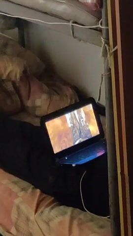 The image captures a person lying on the lower bunk of a bunk bed, likely in a dormitory, barracks, or similar shared living space. The individual, partially covered by a dark blanket or clothing, is engaged in watching content on a small laptop or 2-in-1 device resting on their lap. The device's screen displays a video, which appears to show a performance or concert with bright stage lighting and several figures.

A white cable, possibly a charging cord or headphone wire, extends from the device. The bunk bed features a simple metal frame, around which a white string or rope is tied. A pillow and a patterned blanket with beige and pink hues are visible on the bed. To the right of the frame, a vertical wooden beam or wall section with red and yellow stripes can be seen. The overall lighting in the room appears dim, suggesting it might be evening or nighttime. No legible text is visible in the image.