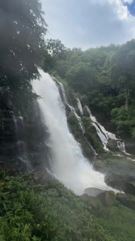 This media file shows a majestic waterfall cascading down a rocky, lush green mountainside. The scene is set in a dense, tropical forest with vibrant green foliage covering the slopes and framing the waterfall. The water is a powerful white torrent, crashing over rocks and creating mist that hangs in the air, contributing to the ethereal atmosphere.

The time of day appears to be daytime, with natural light illuminating the scene. The sky is visible in patches, with hints of clouds, suggesting a partly cloudy or potentially humid day, common in tropical environments like Ban Sop Hat, Thailand.

In the distance, a small group of people can be seen standing on a pathway near the base of the waterfall, offering a sense of scale to the immense natural feature. They appear to be observing the waterfall, possibly enjoying the scenic beauty. The overall mood conveyed is one of awe and the raw power of nature, with the lush greenery and the dynamic water creating a serene yet invigorating environment. There is no visible text in the media file.