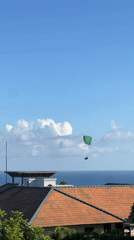 Green-red paraglider spotted soaring over water beneath cloudy skies