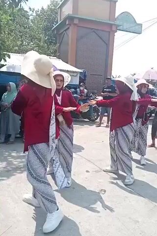The video shows a group of four women in traditional Indonesian attire performing a dance outdoors. They are all wearing maroon tops, white hats, and patterned sarongs. The women are positioned in a semi-circle and appear to be coordinated in their movements. In the background, there are several other people, some on motorcycles, and stalls or temporary structures, suggesting a public gathering or market. The environment is sunny, with a clear sky. The camera is positioned at a medium distance, capturing the entire group of dancers and some of the surrounding activity. The overall atmosphere seems festive.