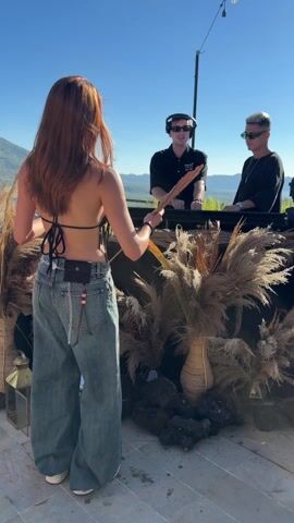 The video shows two male DJs with sunglasses standing behind a table decorated with pampas grass and other decorative elements. One DJ is adjusting the equipment while wearing headphones. The other DJ is making hand gestures. In the foreground, a woman with red hair is holding a golden fan and moving it in time with the music. The background shows a mountain landscape under a clear blue sky. There are string lights overhead.