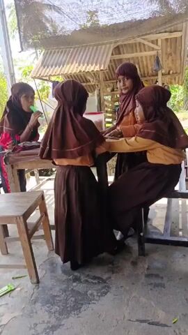 Four young girls wearing brown hijabs and school uniforms are gathered around a table under a corrugated metal roof. One girl is eating a popsicle. The other three girls are interacting around the table, one standing and the others seated. One seated girl briefly stands up and points her finger toward the camera before resuming her seated position. The setting appears to be outdoors, with glimpses of greenery and a simple structure in the background. The ground is a painted gray surface.