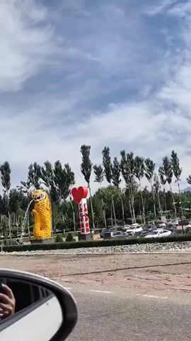 The video shows a street view with a large, yellow owl statue on the left and a red heart sculpture with white stripes on the right. The owl statue appears to be a fountain, with water spraying from its mouth. The statues are surrounded by a line of tall, slender trees. In the background, there is a parking lot with several cars. The sky is partly cloudy. In the foreground, the edge of a road and the side mirror of a vehicle are visible. A white police car with blue and white markings is parked on the right side of the road. The overall scene appears to be an outdoor public space, possibly an entrance to a town or park.