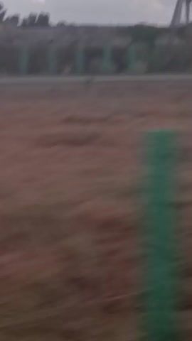 The video shows a grainy, out-of-focus landscape with large industrial cranes in the background. The foreground is blurred, with some green vertical objects visible. The sky is cloudy. There are no people or clear actions depicted.