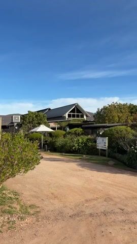 The video shows a large, modern building with a dark roof and a large triangular window on the upper level. The building is surrounded by lush green trees and bushes, with a dirt road in the foreground. The sky is blue with some white clouds. There is a white umbrella and a sign near the building. The camera pans from left to right, showing the building and its surroundings. There are no people visible in the video.