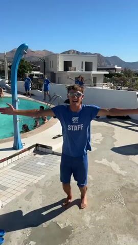 A young man wearing a blue t-shirt with "STAFF" written on it and blue shorts stands barefoot on a concrete surface by a swimming pool. He is wearing sunglasses and performing a dance with his arms. In the background, several other people in blue shirts are in the pool, and some are standing by the pool edge. The scene is outdoors on a sunny day with a clear blue sky and mountains in the distance. The environment appears to be a resort or a facility with a pool.