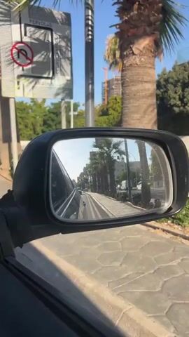 A side mirror of a car reflects a street scene. Palm trees line the road, and buildings are visible in the background. Other cars are also seen on the road, with some moving in the same direction as the car the mirror is attached to. The sky is clear and blue. The car appears to be in motion, as indicated by the blur of the passing scenery.