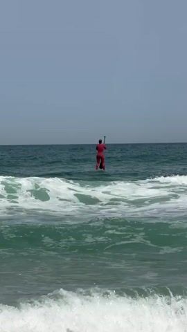 A person in a red wetsuit stands on a paddleboard in the ocean and paddles further out. The water is teal, and some waves are rolling towards the shore. The sky is a pale blue.