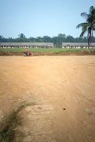 A person rides a motorcycle on a dirt road. In the background, there are buildings and palm trees. A large piece of equipment, possibly construction machinery, is parked to the right of the frame. The scene appears to be an outdoor environment.
