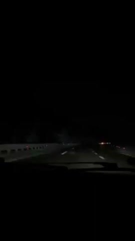 The video depicts a dark, nighttime scene of driving on a highway. The road is marked with white lines and there are guardrails visible on the left side. Headlights from other cars are visible in the distance. The view is from inside a vehicle, looking out the windshield.
