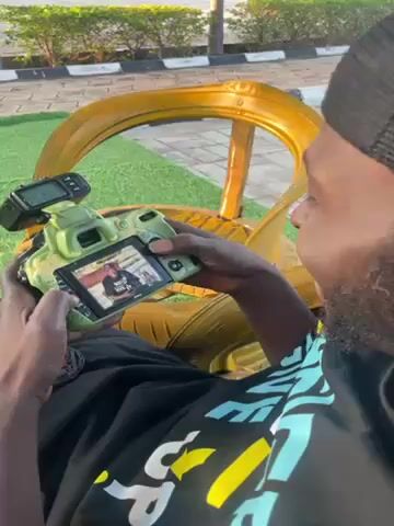 A man is sitting in a gold painted outdoor chair, looking at the LCD screen of a green camouflage patterned DSLR camera that is in his hands. He is smiling, apparently amused by something that he is seeing on the screen. He is wearing a black t-shirt, a black mesh baseball cap with a red element, and blue athletic pants. The man is dark-skinned with a short beard and a mustache. The chair is made of painted tires arranged to resemble a peacock. The scene is outside. There are sidewalks, shrubbery, and palm trees visible in the background.
