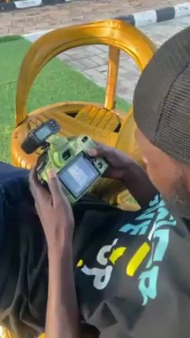 The video shows a person sitting in a gold painted chair. They are wearing a black shirt with white and green graphics on it, a black beanie, and jeans. The person is holding a camera with a green camouflage cover on it. The camera has a flash attachment on top. The person is looking at photos on the camera's display screen. The background includes green grass and a white object.
