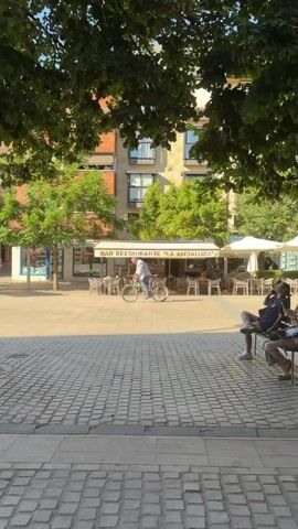 A man rides a bicycle down a paved sidewalk in front of a building with an awning that reads "BAR RESTAURANTE". The man is wearing a checkered shirt and blue jeans. He is riding a black bicycle with disc brakes. Trees and tables and chairs for an outdoor seating area are visible. A woman is sitting at one of the tables, and another person is walking in the background. The sun is shining, creating shadows on the ground.