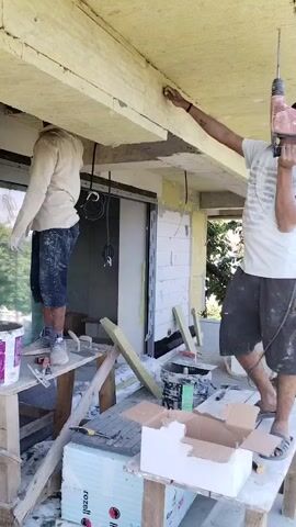 The video shows two men working on the exterior of a building, possibly a balcony or patio area. The man on the left, wearing a light-colored shirt and dark shorts, is standing on a wooden sawhorse and appears to be attaching something to the ceiling, holding a white object in his gloved hands. The man on the right, wearing a white t-shirt and dark shorts, is standing on a similar sawhorse and is holding a red power drill with a long bit, seemingly preparing to drill into the ceiling beam above. Both men are positioned under an overhang, which appears to be made of light-colored insulation material. There are construction materials scattered around, including paint cans, boxes, and pieces of wood or insulation. In the background, another person can be seen on scaffolding, and a third person is partially visible further back, also engaged in work. The environment appears to be a sunny day, with some greenery visible in the background.