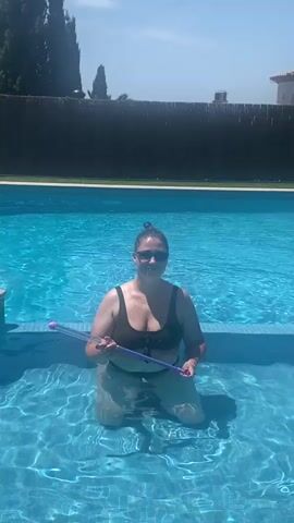 A woman wearing a black bikini and sunglasses is kneeling in a swimming pool. She is holding a purple water gun in her hands. The water gun is a long, thin tube with purple caps on each end. The woman is looking at the camera and smiling. She is in a swimming pool with blue water. There is a black fence in the background, and trees and a house are visible behind the fence. The sky is blue and there are some clouds. The woman raises the water gun and sprays water into the pool. She then lowers the water gun and smiles at the camera again. She extends her arms to her sides.