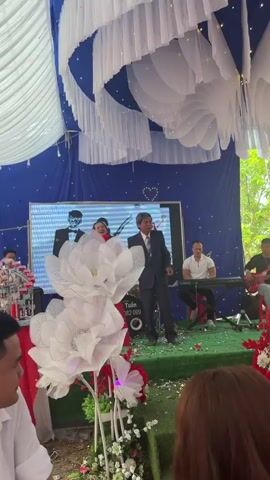 The video captures a celebratory scene, likely an outdoor event or wedding in Tay Son Commune, Vietnam. On a decorated stage with blue and white drapes and a large screen displaying images of a couple, a man in a dark suit with graying hair is standing. A woman in a vibrant pink traditional Vietnamese dress (áo dài) approaches him and hands him a small bouquet of blue flowers. The man smiles and accepts the flowers. To the left, another woman is singing into a microphone, while a man plays a keyboard on the right side of the stage. Large, illuminated white lotus flower decorations adorn the foreground. The stage floor is covered in a green carpet with confetti scattered on it. A drum set is visible behind the woman with the microphone, bearing the text "THUY TUẤN" and the phone number "3 082 099". The keyboard has "YAMAHA" written on it. The overall atmosphere appears joyful and festive.