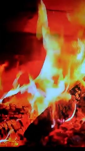 A close-up video shows a fire burning in a fireplace. Logs are visible at the bottom, covered in glowing embers and ash. Flames, predominantly orange and yellow, with some white in the center, leap upwards from the logs. The background is dark, with hints of red and orange suggesting a brick or stone fireplace. The fire appears to be actively burning and casting a warm glow.