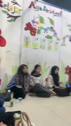 The video shows an indoor scene, possibly a classroom or community space, with several young women sitting on the floor. The room has white walls decorated with colorful drawings, including butterflies, fruits, and trees, and some text in Indonesian. There is a whiteboard or bulletin board mounted on the wall, along with a clock and a fan. In the foreground, a woman is holding a blue cup and a straw. Another woman is looking at her phone. In the background, two more women are sitting on the floor. A red chair and a table with a green top are also visible. The overall impression is a casual gathering.