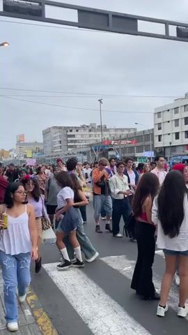 The video shows a street scene with a large crowd of people. Many people appear to be participating in a parade or march. There are buildings on both sides of the street, and an overhead traffic light or sign is visible at the top of the frame. Some individuals are holding flags or banners, and one person is riding a bicycle. The sky is overcast. The people in the crowd are diverse in age and appearance, and many are dressed casually. The overall impression is of a public event or gathering.