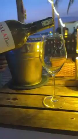The video shows a bottle of DALAT BECO wine being poured into a wine glass on a wooden table. The bottle is dark green, and the label is partially visible. Next to the glass is a metal ice bucket. The scene is illuminated by warm yellow lighting, possibly from a candle or lantern on the table, and there are string lights visible in the background, suggesting an outdoor setting, possibly a beach bar, at dusk. There are blurred figures and structures visible in the background, indicating other patrons and buildings.