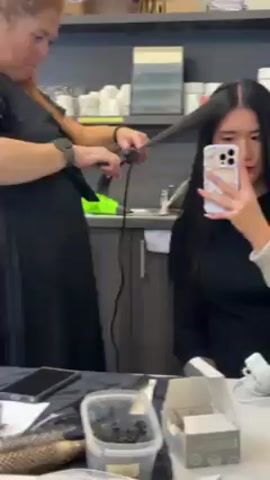 The video shows a woman with red hair wearing a black dress using a hair straightener on a woman with long black hair. The woman being styled is holding a cell phone in front of her face.  The hairstylist is standing on the left, and the client is sitting in front of a mirror. There is a shelf with cups, a green hairdryer, and a gray cabinet behind the hairstylist.  On the counter in front of the client, there is a cell phone, a container of hair clips, and a box.