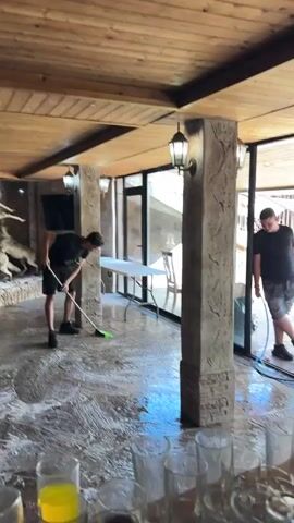 Two young men are cleaning a room with a concrete floor. One man on the left is using a green-bristled broom to push foamy water across the floor. He is wearing a black t-shirt and black shorts with white stripes. The second man on the right is holding a blue hose, which is spraying water. He is wearing a black t-shirt and denim shorts. The room has large glass windows, wooden-paneled ceiling, and decorative stone pillars. There are also stuffed animals, a television screen, a white folding table, and chairs visible in the background. In the foreground, there are several clear drinking glasses filled with yellow liquid.