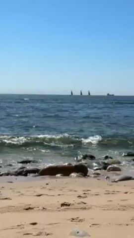 The video shows a sunny beach scene. There is light tan sand on the beach, and several dark rocks are near the water's edge. The ocean is blue with small waves lapping at the shore. In the distance, there are three sailboats with black sails and a larger cargo ship. The sky is clear and blue.