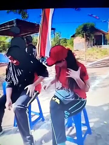 The video shows a group of people participating in an outdoor event, possibly a competition. In the foreground, two women are seated on blue plastic stools. The woman on the left wears a red shirt, black pants with white stripes, and a red baseball cap. She is leaning forward, with her head down and hair covering her face, suggesting she might be performing a task or experiencing discomfort. She has a dark bag slung over her shoulder. The woman on the right wears a purple hijab, a red shirt, and white pants. Her face is adorned with yellow sticky notes, creating a humorous or challenging effect. She appears to be looking forward with a neutral expression. Further to the right, another woman is partially visible, wearing a pink hijab and a pink patterned top. The background reveals a race track or path, with flags and banners, including red and white ones, suggesting a festive occasion or a national celebration. There are buildings and trees in the distance, and the sky is blue. The overall scene conveys a sense of community and lighthearted competition.