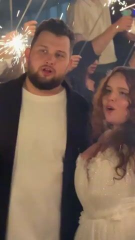 The video shows a group of people celebrating outdoors at night, holding sparklers. In the foreground, a man with a beard and a white t-shirt under a black jacket is looking to his left. Next to him, a woman in a white dress with curly hair is looking to her left and talking. Other people are visible in the background, also holding sparklers and appearing to be in conversation. The sparklers are creating bright bursts of light. The overall atmosphere is festive.